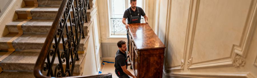 Deux débarrasseurs en pied manœuvrant buffet ancien dans escalier étroit immeuble haussmannien parisien