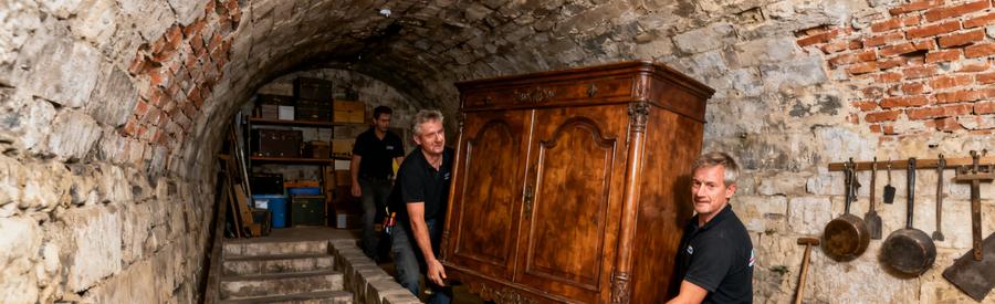 Des débarras professionnels manœuvrant un meuble dans une cave voûtée de Verdun, avec des escaliers étroits et un décor historique.